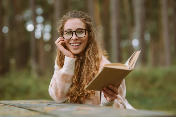 幸せな若い女性は美しい森の中の本とテーブルに座っている 美しい女性は森の読書本で新鮮な空気を楽しんでいます リラクゼーションの概念 自然との孤独 ライフスタイル — ストック写真