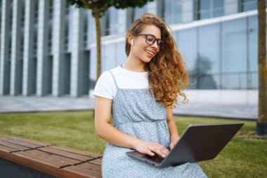 Gülümseyen serbest çalışan kadın dizüstü bilgisayarı ve kablosuz kulaklıkla şehir caddesinde oturuyor. Laptoplu genç bir kadın dışarıda çalışıyor. Eğitim kavramı, İnternet, iş