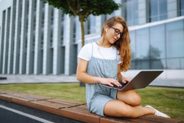 Gülümseyen serbest çalışan kadın dizüstü bilgisayarı ve kablosuz kulaklıkla şehir caddesinde oturuyor. Laptoplu genç bir kadın dışarıda çalışıyor. Eğitim kavramı, İnternet, iş