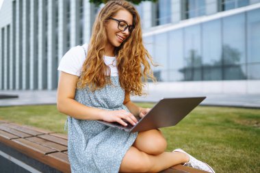 Gülümseyen serbest çalışan kadın dizüstü bilgisayarı ve kablosuz kulaklıkla şehir caddesinde oturuyor. Laptoplu genç bir kadın dışarıda çalışıyor. Eğitim kavramı, İnternet, iş