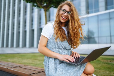 Gülümseyen serbest çalışan kadın dizüstü bilgisayarı ve kablosuz kulaklıkla şehir caddesinde oturuyor. Laptoplu genç bir kadın dışarıda çalışıyor. Eğitim kavramı, İnternet, iş