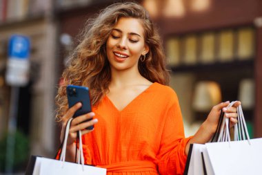 Alışveriş yapan kadın. Şehirdeki alışveriş çantalı genç bir kadın internetten alışveriş yapıyor. Gülümseyen turistler Avrupa sokaklarında yürürken güneşli havanın keyfini çıkarırlar. Çevrimiçi alışveriş kavramı.