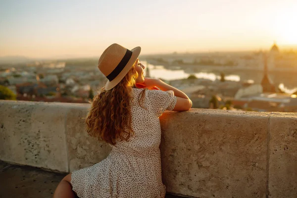 stock image Beautiful woman in hat enjoys stunning view of the city at dawn. Back view. Tourist is looking at landscape of city from height. Travel in Europe.