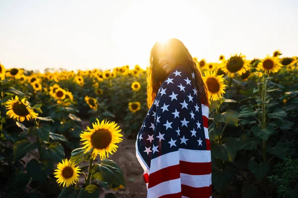 独立日 7月4日日落时分 美丽的年轻女子在向日葵地里举着美国国旗 美丽的日落自由 爱国假日 — 图库照片