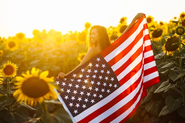 独立日 7月4日日落时分 美丽的年轻女子在向日葵地里举着美国国旗 美丽的日落自由 爱国假日 — 图库照片