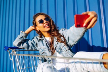 Selfie zamanı. Genç bir kadın cep telefonuyla alışveriş arabasında eğleniyor, selfie çekiyor. Alışveriş zamanı, gençlik konsepti, tatil. 