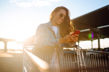 Selfie zamanı. Genç bir kadın cep telefonuyla alışveriş arabasında eğleniyor, selfie çekiyor. Alışveriş zamanı, gençlik konsepti, tatil. 