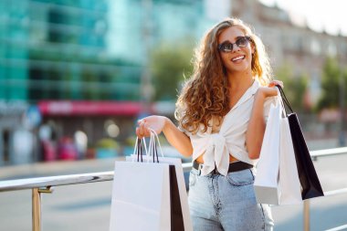 Alışveriş çantalı mutlu bir kadın güneşli bir sokakta yürüyor. Yaşam tarzı, alım, satım, tüketim kavramı.