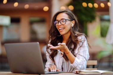Kablosuz kulaklıklı genç bir kadın dizüstü bilgisayardan arıyor, webcam ile konuşuyor, modern kafede video konferansı yapıyor. Eğitim, iş, blog veya serbest çalışma kavramı.
