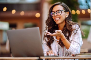 Kablosuz kulaklıklı genç bir kadın dizüstü bilgisayardan arıyor, webcam ile konuşuyor, modern kafede video konferansı yapıyor. Eğitim, iş, blog veya serbest çalışma kavramı.
