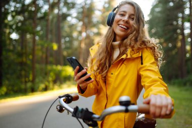 Kulaklıklı ve akıllı telefonlu mutlu bir kadın güneşli bir parkta müzik dinleyerek bisiklete biniyor. Aktif yaşam tarzı, tatil, dinlenme.