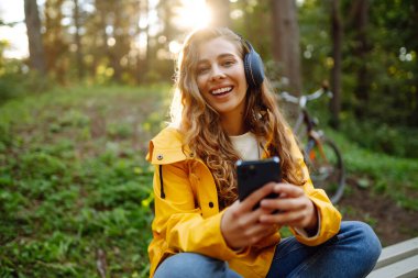 Kulaklıklı ve akıllı telefonlu mutlu bir kadın güneşli bir parkta müzik dinleyerek bisiklete biniyor. Aktif yaşam tarzı, tatil, dinlenme.