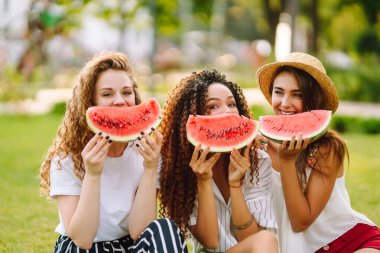 Üç güzel genç kız birlikte eğleniyor ve sıcak bir günde karpuz yiyorlar. Yaz, yaşam tarzı, seyahat, doğa ve tatil kavramı.