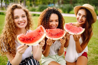 Üç güzel genç kız birlikte eğleniyor ve sıcak bir günde karpuz yiyorlar. Yaz, yaşam tarzı, seyahat, doğa ve tatil kavramı.