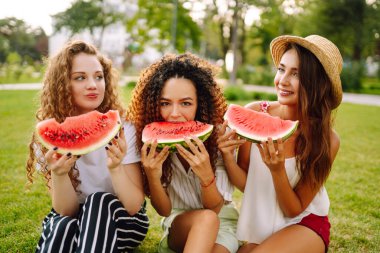 Üç güzel genç kız birlikte eğleniyor ve sıcak bir günde karpuz yiyorlar. Yaz, yaşam tarzı, seyahat, doğa ve tatil kavramı.