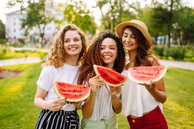 Üç güzel genç kız birlikte eğleniyor ve sıcak bir günde karpuz yiyorlar. Yaz, yaşam tarzı, seyahat, doğa ve tatil kavramı.