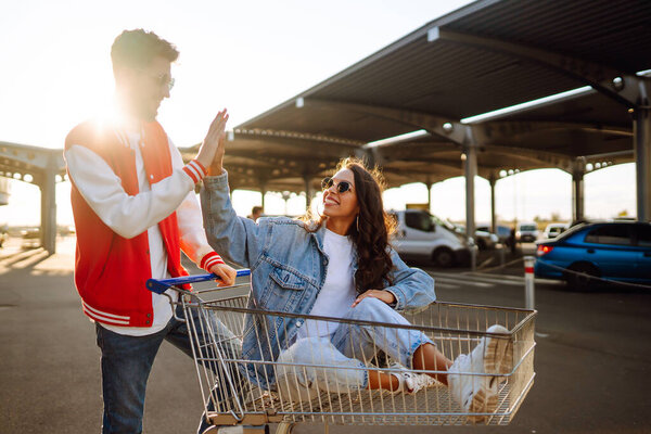 Stylish man and woman having fun and riding shopping cart. Black friday. Consumerism, sale, discounts, lifestyle concept.