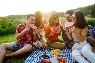 Bir grup arkadaş birlikte eğlenir ve sıcak yaz gününde karpuz yerler. İnsanlar, yaşam tarzı, seyahat, doğa ve tatil kavramı. 