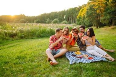 Bir grup arkadaş birlikte eğlenir ve sıcak yaz gününde karpuz yerler. İnsanlar, yaşam tarzı, seyahat, doğa ve tatil kavramı. 