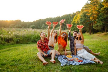 Bir grup arkadaş birlikte eğlenir ve sıcak yaz gününde karpuz yerler. İnsanlar, yaşam tarzı, seyahat, doğa ve tatil kavramı. 