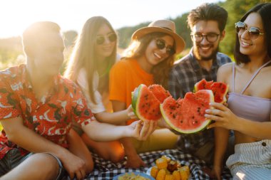 Bir grup arkadaş birlikte eğlenir ve sıcak yaz gününde karpuz yerler. İnsanlar, yaşam tarzı, seyahat, doğa ve tatil kavramı. 