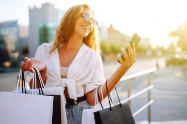 Güneşli bir sokakta telefonla alışveriş yapan şık genç bir kadın. Çevrimiçi alışveriş, satış, tüketim kavramı. Etkin yaşam biçimi.