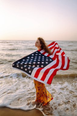 Gün batımında kumsalda Amerikan bayrağı taşıyan genç bir kadın. 4 Temmuz. Bağımsızlık Günü. Vatansever bayram.