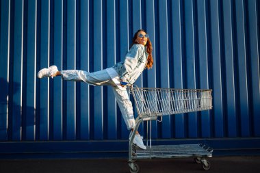 Alışveriş arabalı genç bir kadın renk duvarının yanında eğleniyor. Gülümseyen Afrikalı kadın alışverişe çıkıyor.. 