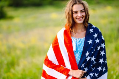 Çiçekli çayırda Amerikan bayrağı taşıyan genç bir kadın. 4 Temmuz. Bağımsızlık Günü. Vatansever bayram. ABD bayrağı rüzgarda dalgalanıyor.