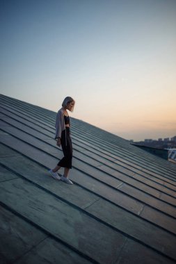 Çatıda fotoğraf çekimi. Gün batımında çatıda poz veren genç bir kadın. İnsanlar, yaşam tarzı, rahatlama konsepti.