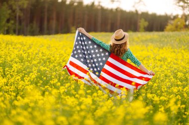 Çiçekli çayırda Amerikan bayrağı taşıyan genç bir kadın. 4 Temmuz. Bağımsızlık Günü. Vatansever bayram. ABD bayrağı rüzgarda dalgalanıyor.