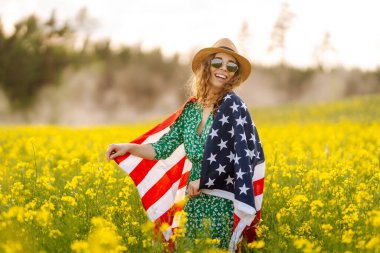 Çiçekli çayırda Amerikan bayrağı taşıyan genç bir kadın. 4 Temmuz. Bağımsızlık Günü. Vatansever bayram. ABD bayrağı rüzgarda dalgalanıyor.