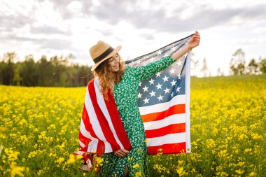 Çiçekli çayırda Amerikan bayrağı taşıyan genç bir kadın. 4 Temmuz. Bağımsızlık Günü. Vatansever bayram. ABD bayrağı rüzgarda dalgalanıyor.