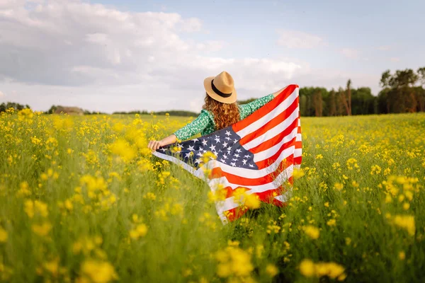 Young woman with american flag on blooming meadow. 4th of July. Independence Day. Patriotic holiday. USA flag fluttering in the wind.