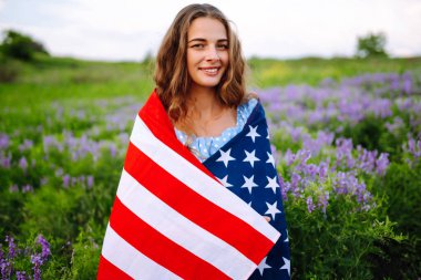 Çiçekli çayırlarda Amerikan bayrağı sallayan genç bir kadın. 4 Temmuz. Bağımsızlık Günü. Vatansever bayram. ABD bayrağı rüzgarda dalgalanıyor.