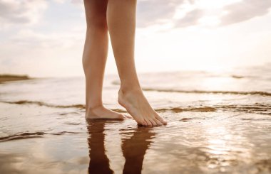 Kadın bacağı, gün batımında kumsalda yürürken yakınlaşıyor ve dinleniyor. Seksi, zayıf ve bronzlaşmış bacaklar. Yaz tatili.