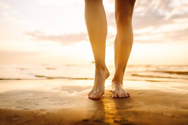 Kadın bacağı, gün batımında kumsalda yürürken yakınlaşıyor ve dinleniyor. Seksi, zayıf ve bronzlaşmış bacaklar. Yaz tatili.
