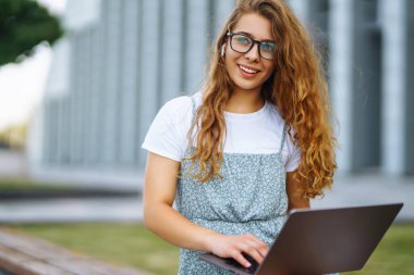Parkta oturan, dizüstü bilgisayar kullanan ve daktilo yazan genç bir kadın. Eğitim kavramı, çevrimiçi, serbest, iş.