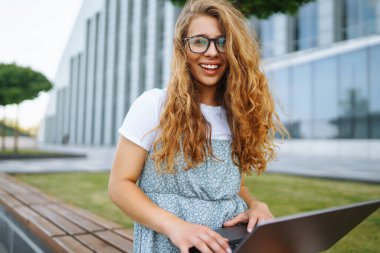 Parkta oturan, dizüstü bilgisayar kullanan ve daktilo yazan genç bir kadın. Eğitim kavramı, çevrimiçi, serbest, iş.