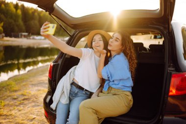 İki genç kadın yolculuk sırasında arabada selfie çekiyor. Yaşam tarzı, seyahat, turizm, doğa, aktif yaşam.