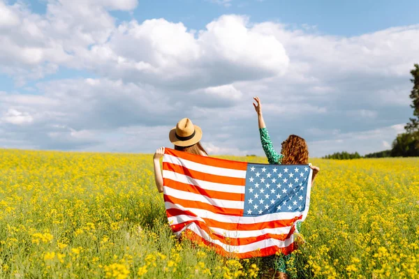 Two Young woman with american flag on blooming meadow. 4th of July. Independence Day. Patriotic holiday. USA flag fluttering in the wind.