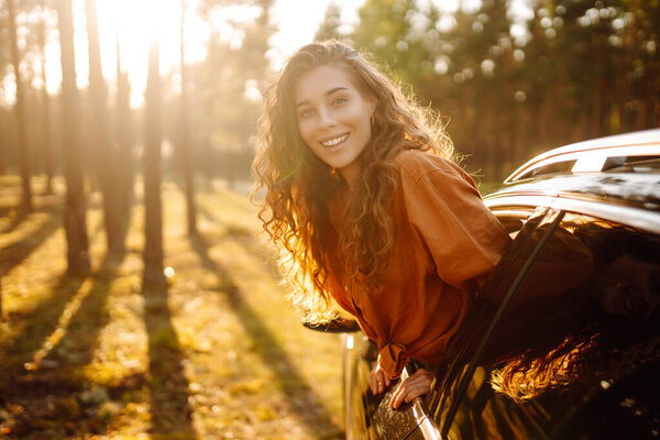 Beautiful traveler woman is resting and enjoying the sunset in the car. Happy woman on a summer trip, travels leaning out of the car window. Active lifestyle, tourism. Travel concept.
