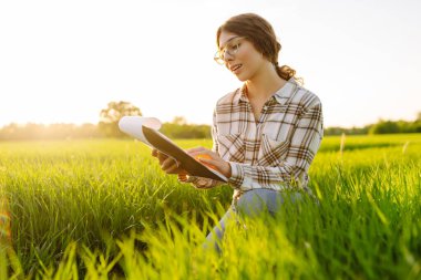 Genç dişi çiftçi, tarladaki genç buğdayla birlikte bir tabletle çalışıyor. Çiftçi buğday filizlerine dokunur, büyümeyi kontrol eder. Çiftlik gelişimi ve bahçıvanlık kavramı.