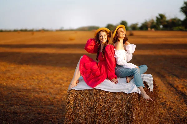 Beautiful two women have fun in a haystack at sunset. Stylish girlfriends in hats have fun on the field. The concept of nature, lifestyle and recreation.