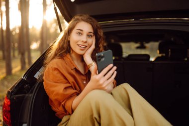 Güzel bir kadın ormanda bir arabanın bagajında oturur ve akıllı telefon kullanır. Arabayla seyahat etme kavramı, aktif yaşam tarzı..