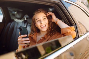 Bir arabanın arka koltuğunda otururken akıllı telefon kullanan gülümseyen bir kadın. Genç kadın arabada postaları, mesajları, blogları kontrol ediyor. İş, teknoloji kavramı.