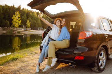 Gülümseyen genç bir kadın seyahat ederken arabanın arka koltuğunda eğlenir. Kız arkadaşlar arabada oturup güneşli havanın tadını çıkarırken selfie çeker, güler ve dinlenirler. Etkin yaşam biçimi.