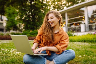 Güzel kadın serbest yazar elinde dizüstü bilgisayarla yeşil bir çayırda oturuyor. Online eğitim. Güneşin tadını çıkaran mutlu bir kadın. Bağımsız kavram.