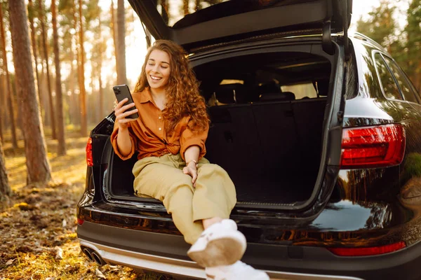 美丽的女人坐在森林里一辆汽车的后备箱里 用的是智能手机 驾车旅行的概念 积极的生活方式 — 图库照片