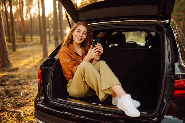 美丽的女人坐在森林里一辆汽车的后备箱里 用的是智能手机 驾车旅行的概念 积极的生活方式 — 图库照片